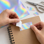 Ejemplo ilustrativo de uso del papel adhesivo holográfico: retirar el respaldo y aplicar un recorte en la cubierta de un cuaderno. Escena generada por IA, no fotografía oficial ni prueba de rendimiento. Cuaderno y tijeras no incluidos.